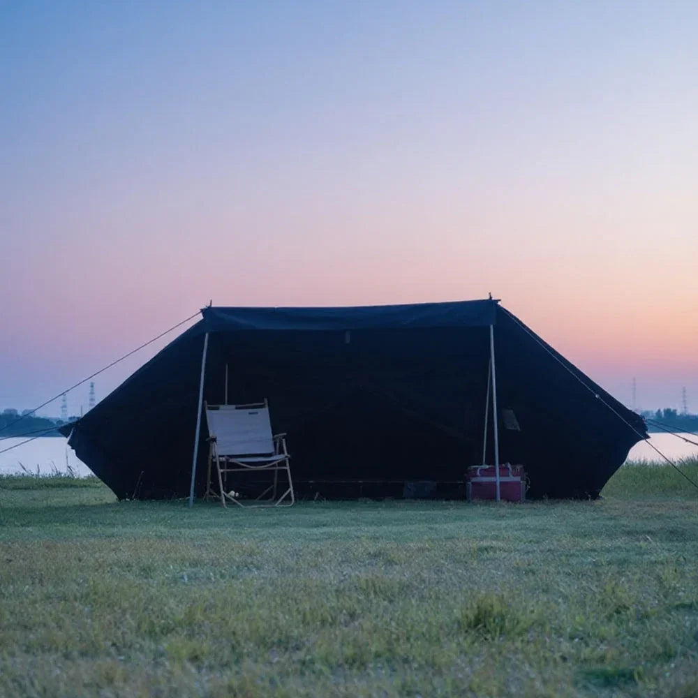 Large black camping tent with a folding chair on grassy field by a river at sunset