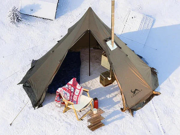 Large canvas camping tent with stove jack set up in snowy outdoor setting, chair and table outside.
