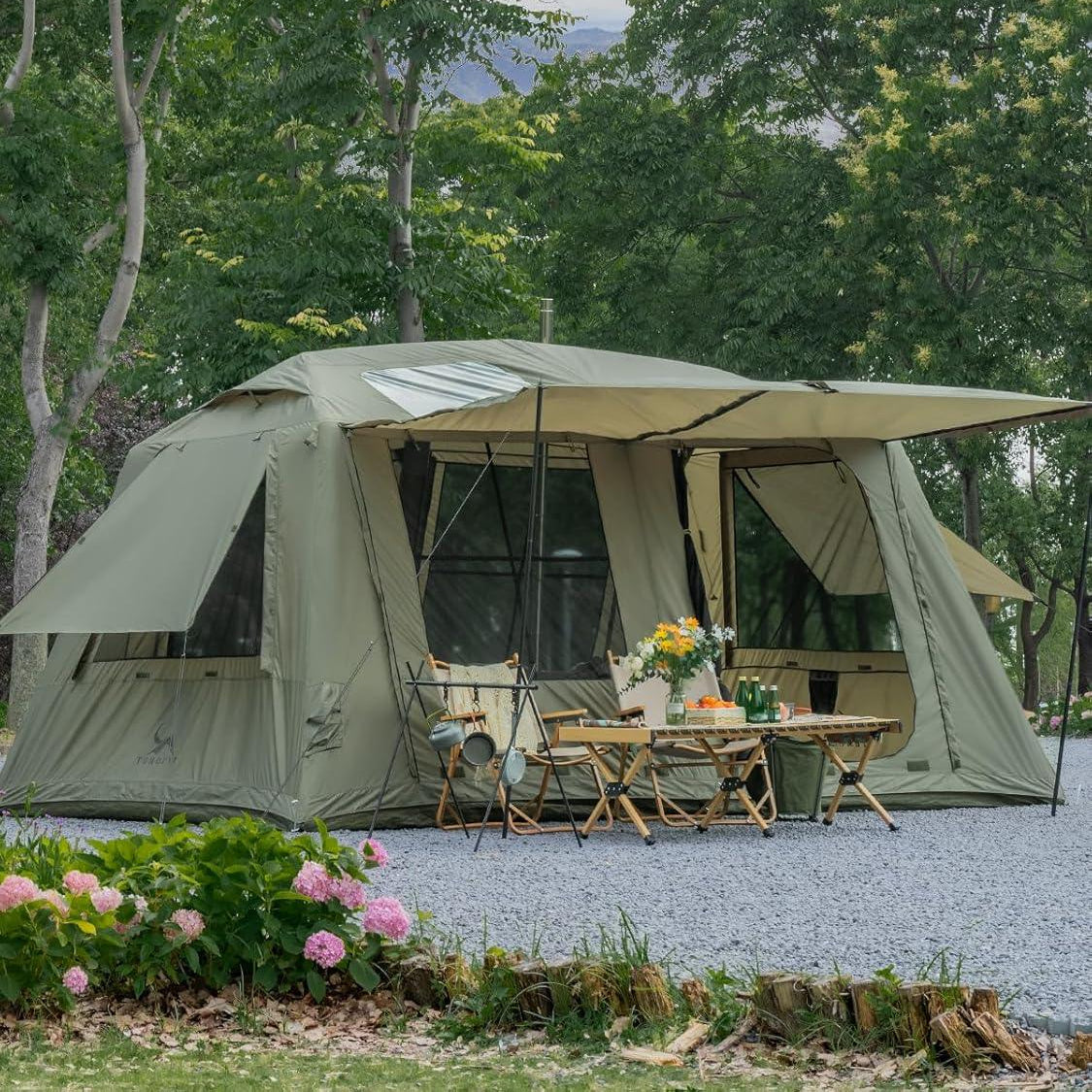 Large green camping tent with open awning, outdoor dining setup, trees and flowers nearby